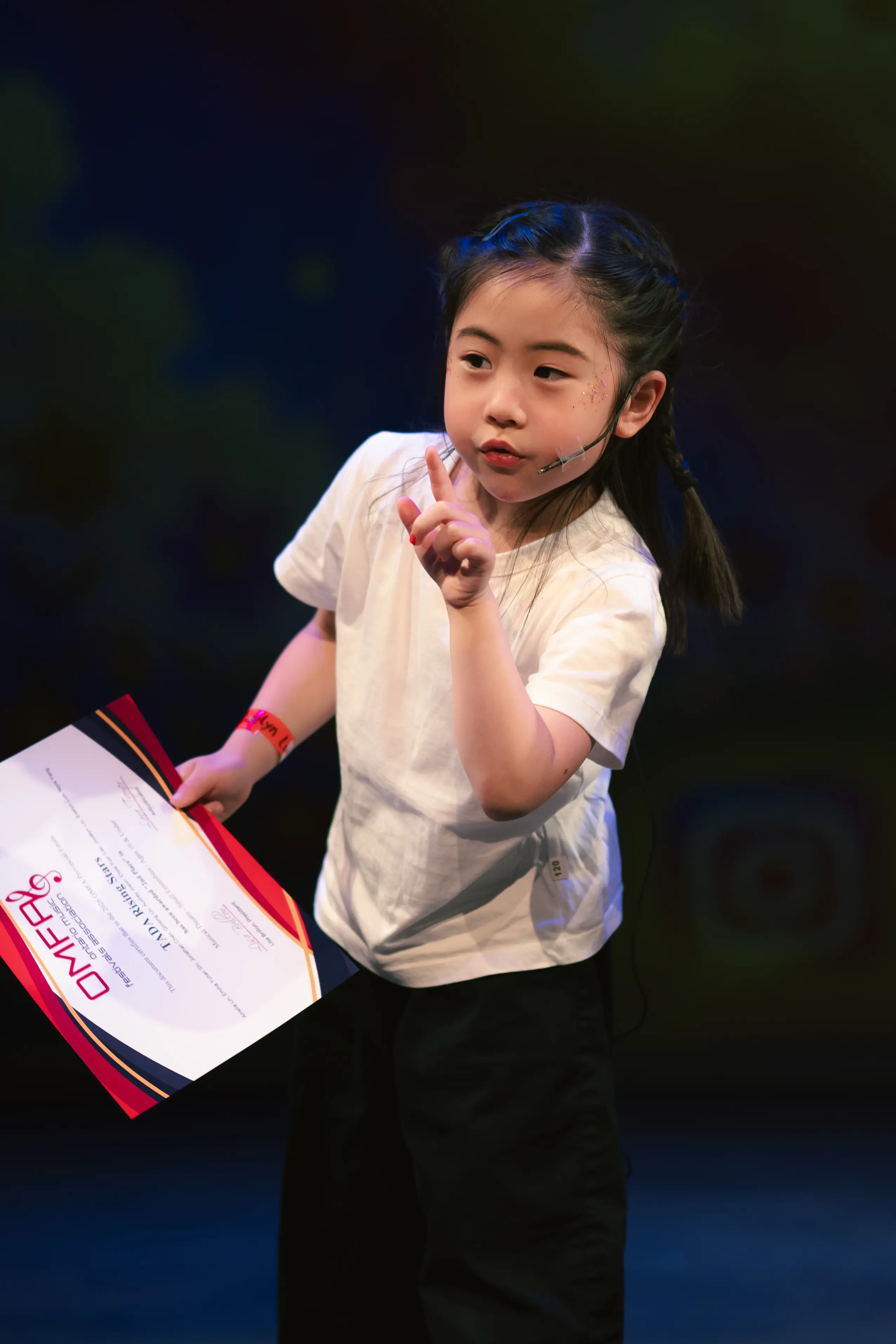 A cute young TADA performer holding a provincial achievement certificate, pointing to the audience.