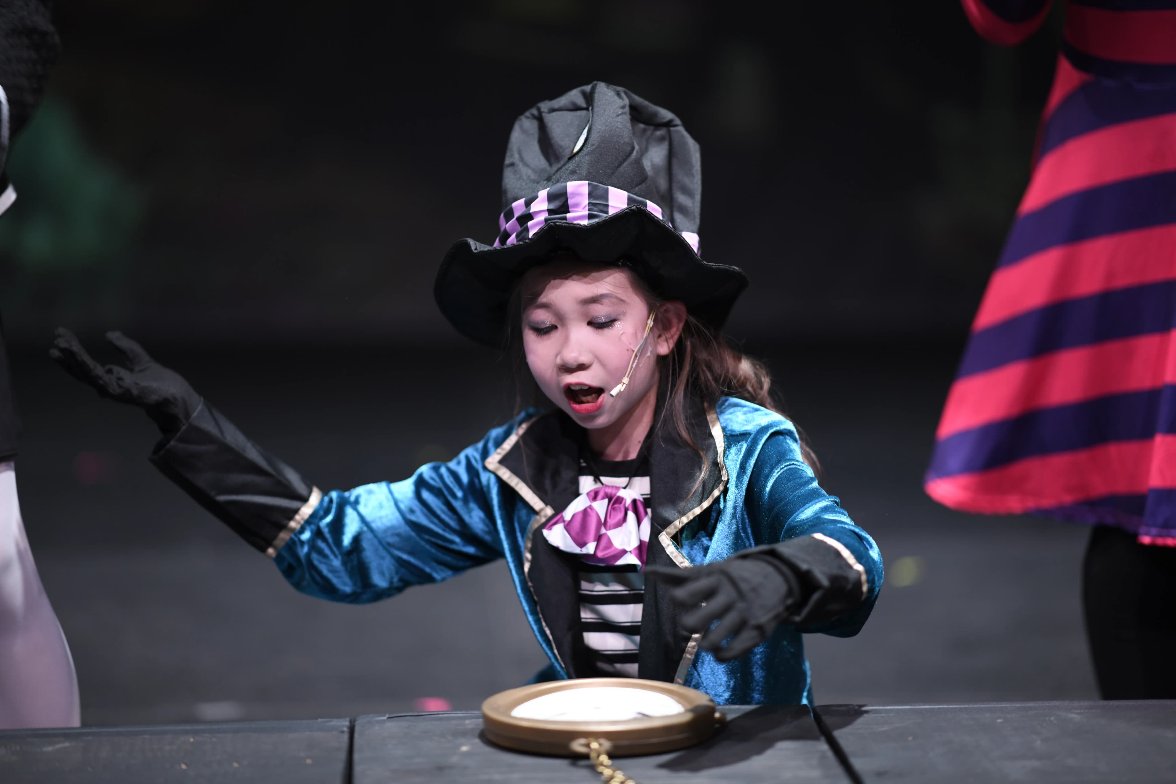 A young performer in a top hat studies a pocket watch onstage.