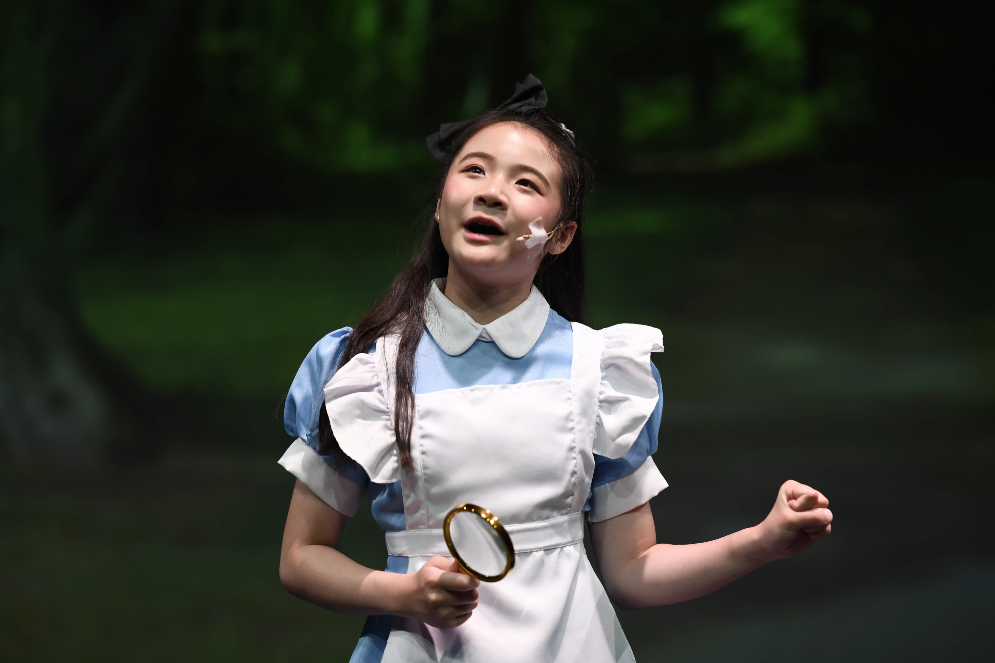 A young performer, in an Alice costume sings on stage with a magnifying glass.