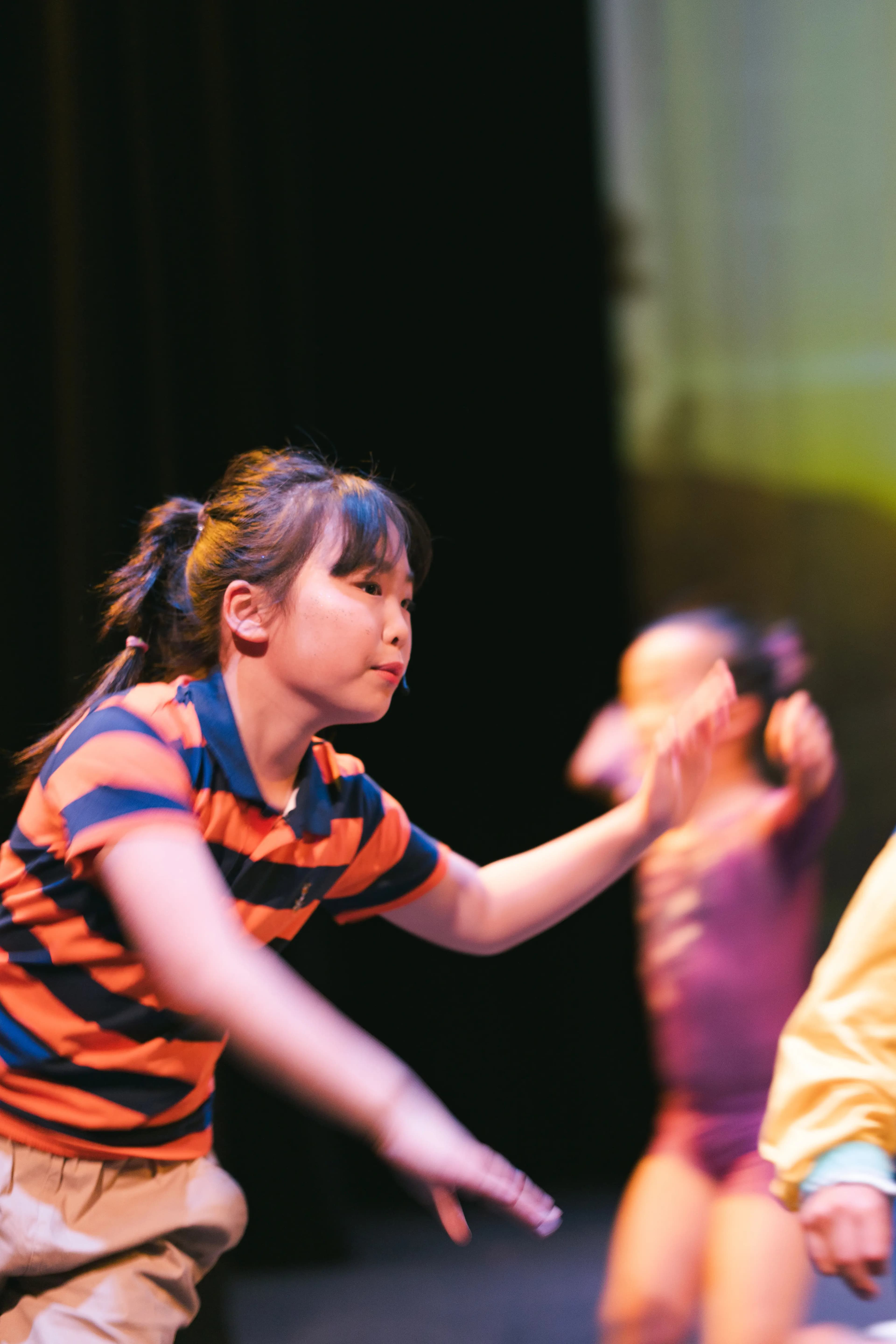 A young performer dances onstage with other students blurred in motion behind her.