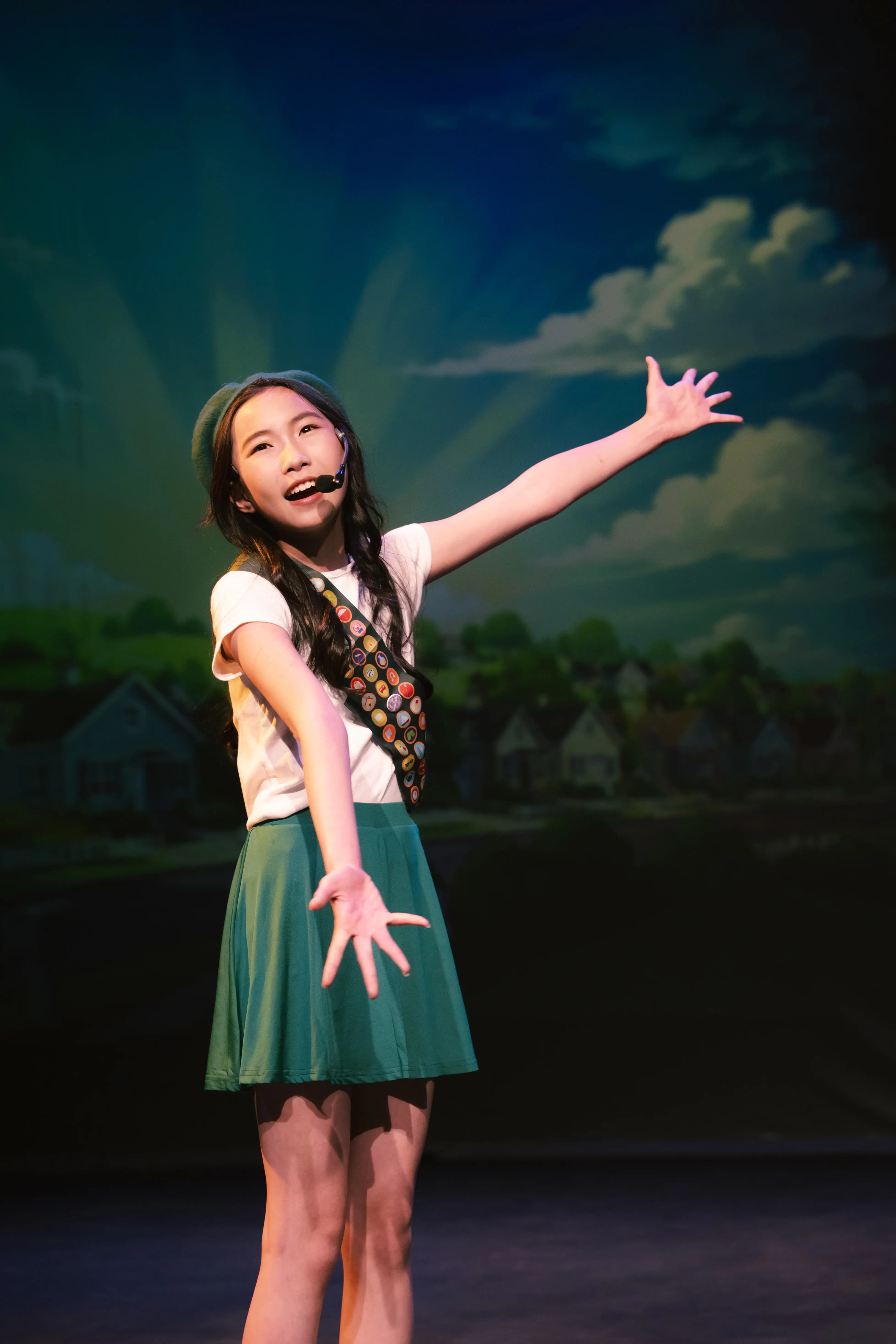 A performer in a Girl Scout costume sings with one arm outstretched.
