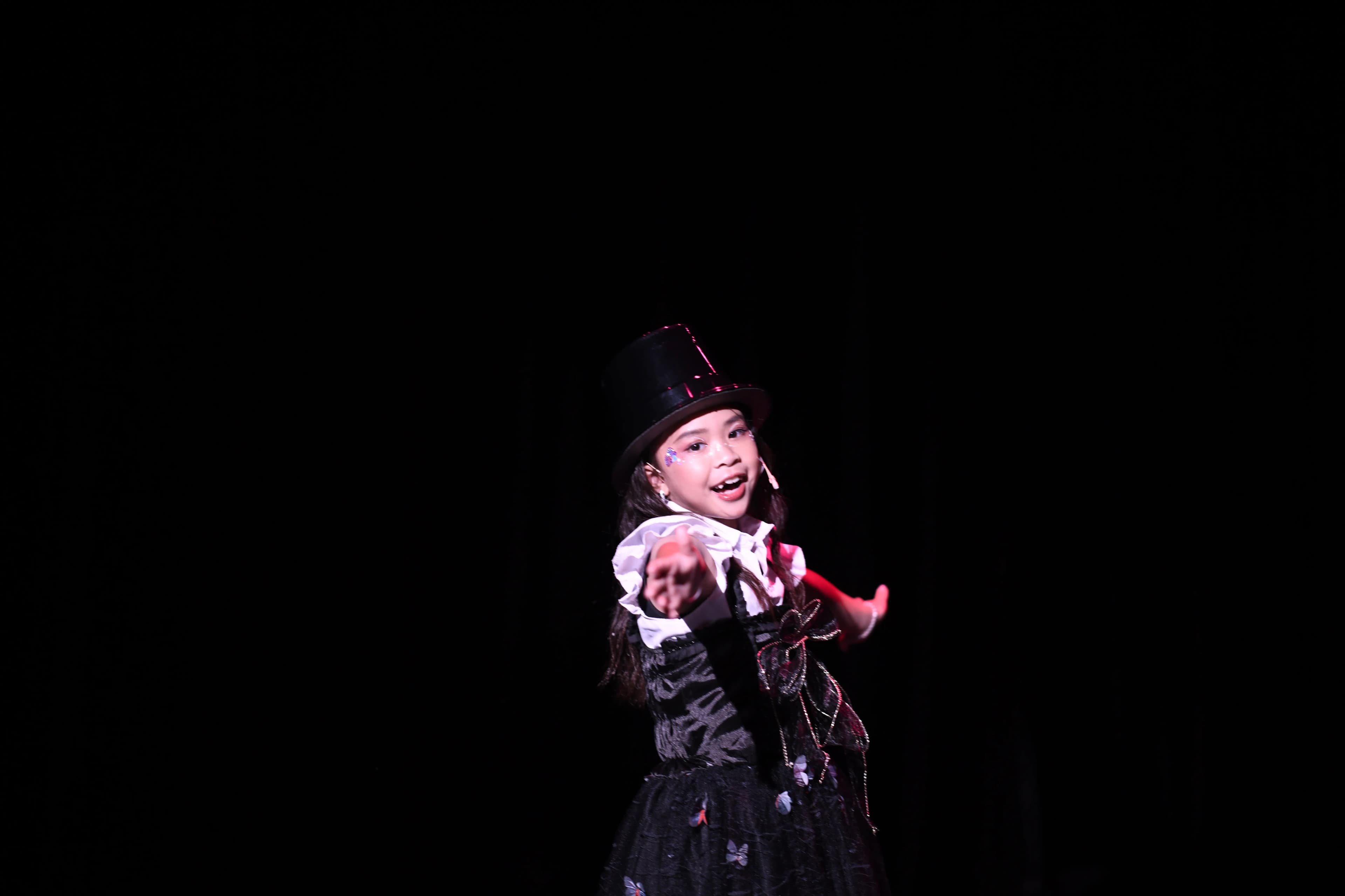 A soloist performs on a black stage in a tophat, pointing and smiling directly into the camera.