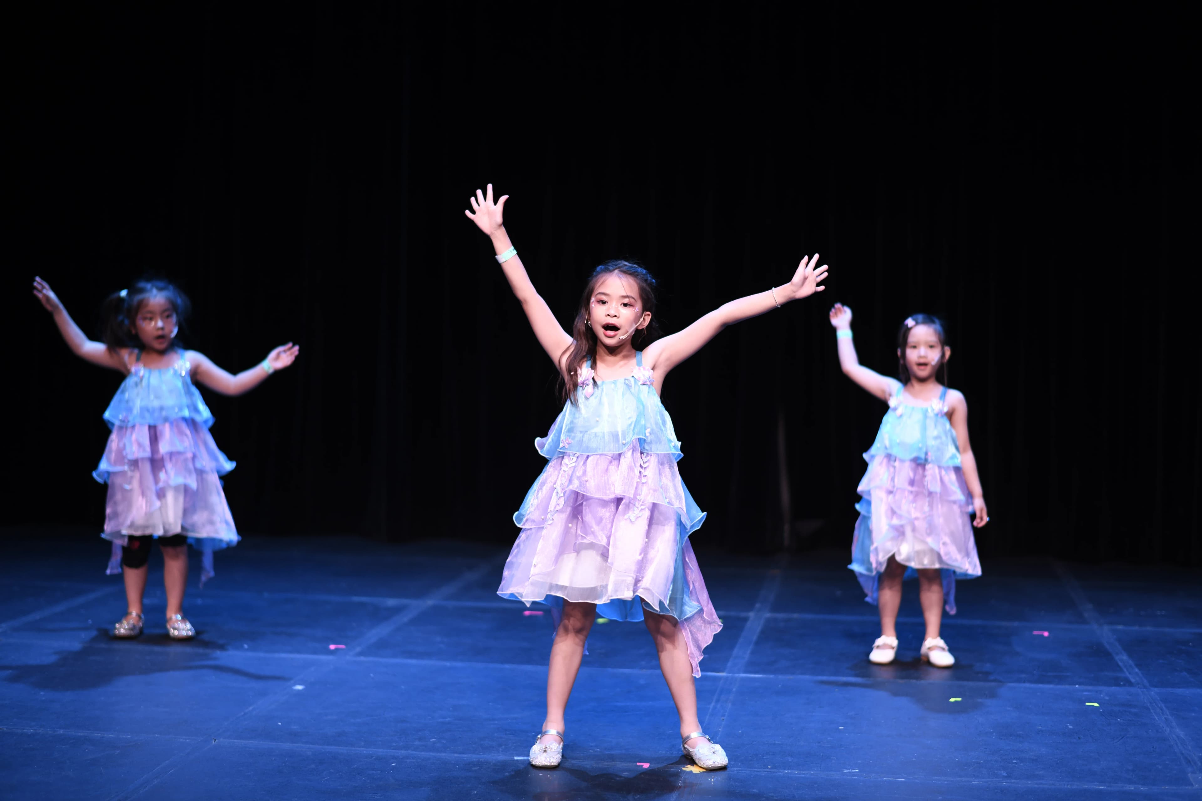 A young singer in a blue-and-purple costume raises her arms onstage.
