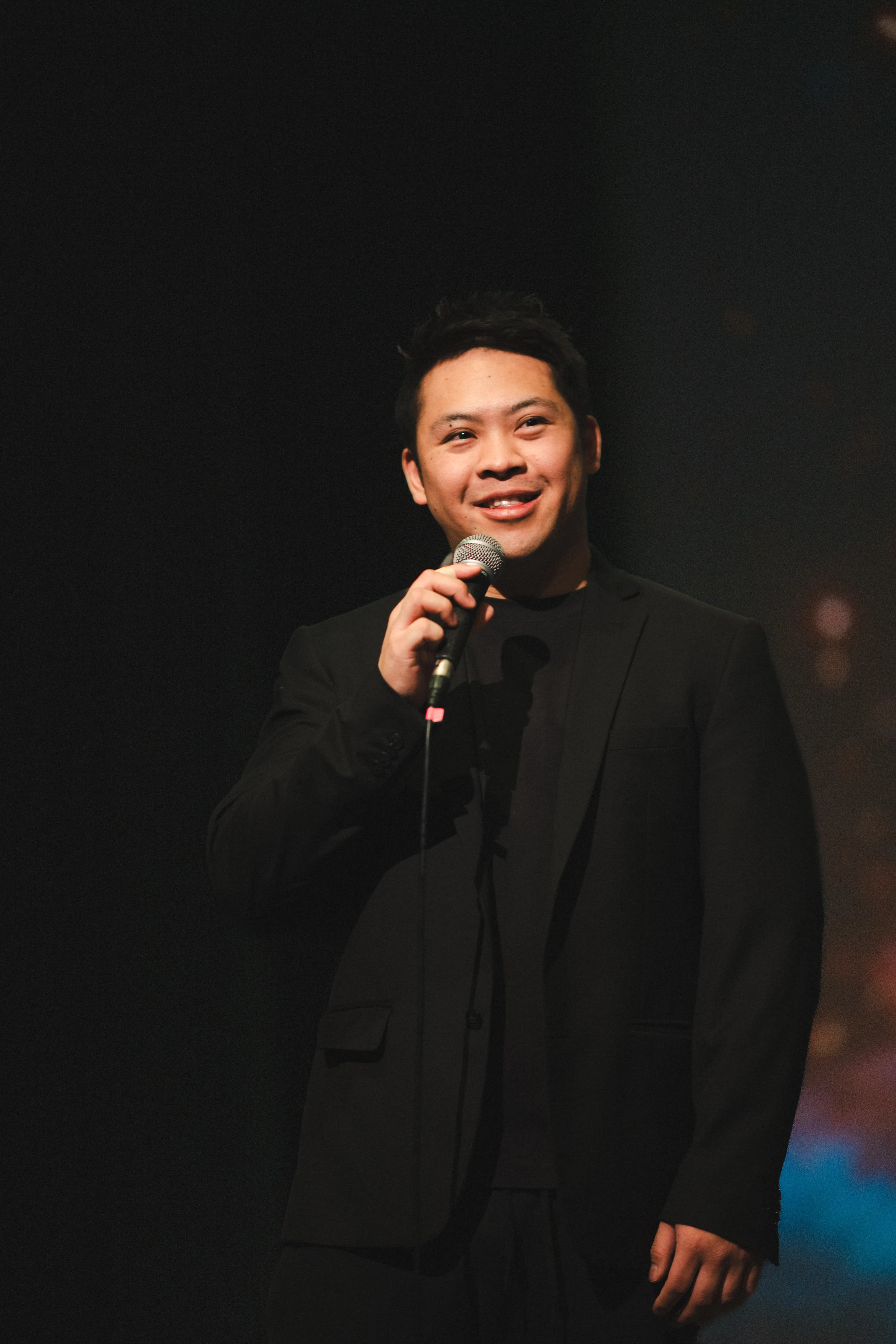 A professional headshot of a teacher, holding a microphone, wearing a black suit.